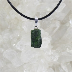May include: A dark green crystal pendant on a black leather cord. The pendant is a raw, unpolished stone with a natural shape.