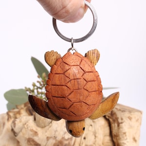 Könnte beinhalten: Ein Holz-Schildkröten-Schlüsselanhänger mit einem silbernen Schlüsselring. Die Schildkröte ist aus hellem und dunklem braunem Holz geschnitzt, mit detaillierten Panzer- und Flossenmustern. Der Schlüsselanhänger vor weißem Hintergrund.