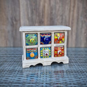Könnte beinhalten: Ein weißer Holzschrank mit sechs Schubladen. Jede Schublade hat ein buntes, handgemaltes Design mit geometrischen Mustern und leuchtenden Farben wie Rot, Blau, Grün, Gelb und Orange.