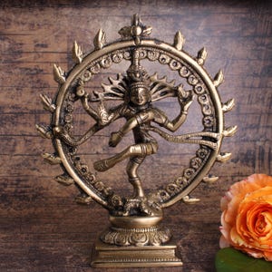Könnte beinhalten: Eine bronzefarbene Statue der hinduistischen Gottheit Shiva in einer Tanzpose, umrahmt von einem detaillierten kreisförmigen Heiligenschein. Die Statue steht vor einem hölzernen Hintergrund, mit einer Rose in der unteren rechten Ecke. Die Statue ist etwa 20 cm hoch.