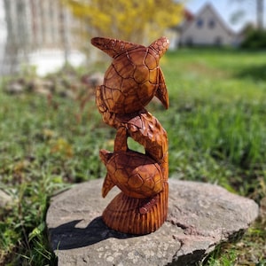 Può includere: Una scultura in legno di tre tartarughe impilate l'una sull'altra. Le tartarughe sono scolpite con dettagliati motivi a guscio e sono di colore marrone caldo. La scultura è posta su una pietra in un ambiente naturale all'aperto.