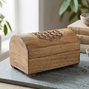 Op de afbeelding: Een rechthoekige houten doos met een gebogen, scharnierend deksel voorzien van een uitgesneden geometrisch patroon op de bovenzijde. De doos heeft een natuurlijke bruine houtnerfafwerking en rust op vier kleine, vierkante voetjes.