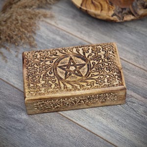 Op de afbeelding: Een rechthoekige houten kist in lichtbruin met ingewikkelde bloemensnijwerk en een centraal pentagramontwerp. De gedetailleerde snijwerken creëren een textuur. De kist wordt waarschijnlijk gebruikt voor opslag.