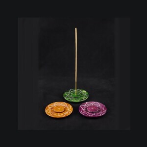 Può includere: Tre porta incensi decorativi a forma di fiore di loto. I supporti sono in pietra e sono dipinti in verde, arancione e viola. Un bastoncino di incenso in legno è in piedi nel supporto verde.