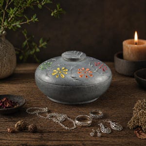 Op de afbeelding: Een grijze, ovale sieradendoos met een deksel, versierd met kleurrijke bloemmotieven. De doos staat op een houten oppervlak met sieraden, een kaars en decoratieve elementen. De bloemen zijn geel, oranje en groen.