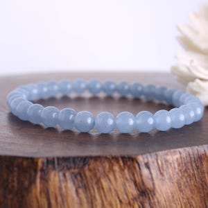 Peut inclure: Un bracelet de perles bleu clair. Le bracelet est composé de perles rondes et polies enfilées ensemble. Les perles ont une couleur bleu pastel douce. Le bracelet est présenté sur une surface en bois.