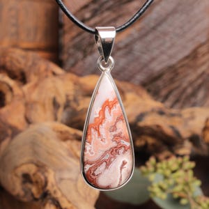 Könnte beinhalten: Ein tropfenförmiger Anhänger mit silberfarbener Öse und einem gemusterten Stein. Der Stein weist eine Mischung aus Farben auf, darunter Weiß, Orange und Braun, mit Marmoreffekt. Der Anhänger hängt an einem schwarzen Band.