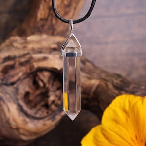 May include: A clear quartz crystal pendant on a black leather cord. The crystal is faceted and has a silver setting. The pendant is hanging against a wooden background.