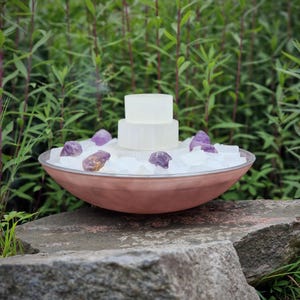 Peut inclure: Un bol cuivré rempli de cristaux blancs et de pierres d'améthyste. Une structure rectangulaire blanche à deux niveaux est placée au centre. Le bol est posé sur une grosse pierre grise, avec un feuillage vert en arrière-plan.