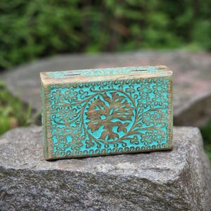 Könnte beinhalten: Eine rechteckige Holzbox mit türkisfarbenem Finish und aufwendigen Blumen- und Baumgravuren. Die Box steht auf einer grauen Steinoberfläche und zeigt die detaillierte Handwerkskunst und natürliche Ästhetik.