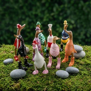 Könnte beinhalten: Eine Sammlung skurriler Holzfiguren von Enten, jede in farbenfrohen Outfits und Hüten. Die Enten sind auf einem Bett aus grünem Moos drapiert, mit kleinen Steinen darum herum. Die Enten tragen Stiefel und Kleider.