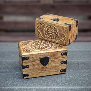 Könnte beinhalten: Zwei Holzkisten mit aufwendig geschnitzten Designs und schwarzen Metallverzierungen. Die obere Kiste hat ein Blumenmuster und die untere Kiste ein Sternmuster.