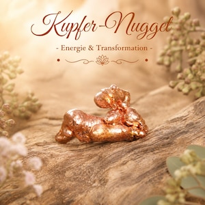 May include: A copper nugget with the text "Kupfer-Nugget - Energie & Transformation" in a decorative font. The nugget is a reddish-brown color and lies on a piece of wood. The background is blurred with hints of greenery.