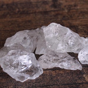May include: A pile of clear quartz crystals on a brown wooden surface. The crystals are various sizes and shapes, with some having a slightly cloudy appearance.