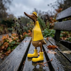 Könnte beinhalten: Eine Entenfigur aus Holz mit natürlicher brauner Oberfläche, die einen leuchtend gelben Regenmantel, einen passenden gelben Regenhut und gelbe Gummistiefel trägt. Die Ente steht im Profil nach links gerichtet und hat eine glatte, geschnitzte Textur.