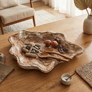 Könnte beinhalten: Eine große, unregelmäßig geformte Holzservierschale mit hellbraunem und weißem Marmor-Finish. Die Schale enthält kleine Steine, zwei Holzlöffel und zwei kleine Granatäpfel. Die Schale steht auf einem Holztisch.