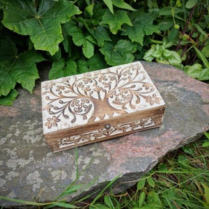 Könnte beinhalten: Eine rechteckige Holzbox mit einem geschnitzten Lebensbaum-Design auf dem Deckel. Die Box ist hellbraun mit weißem Hintergrund und floralen Akzenten. Sie hat einen kleinen Metallverschluss und liegt auf einem großen Stein.