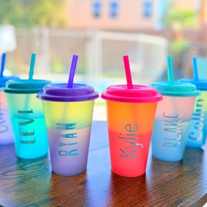 May include: A collection of six color-changing tumblers with lids and straws. Each tumbler has a different color lid and straw, and a name printed on the side. The tumblers are arranged on a wooden table.