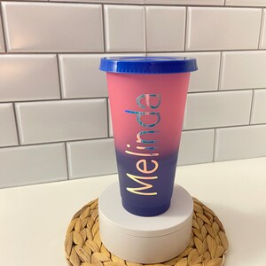 May include: A pink and blue ombre reusable cup with a blue lid. The cup has the name "Melina" written on it in gold lettering.