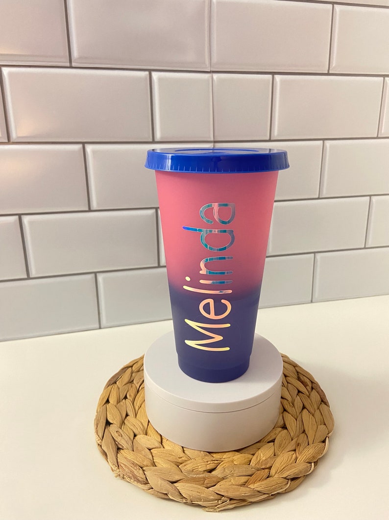 May include: A pink and blue ombre reusable cup with a blue lid. The cup has the name "Melina" written on it in gold lettering.