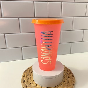 May include: A pink reusable cup with an orange lid and the name "Samantha" written on the side in a silver holographic font. The cup is sitting on a woven straw coaster.