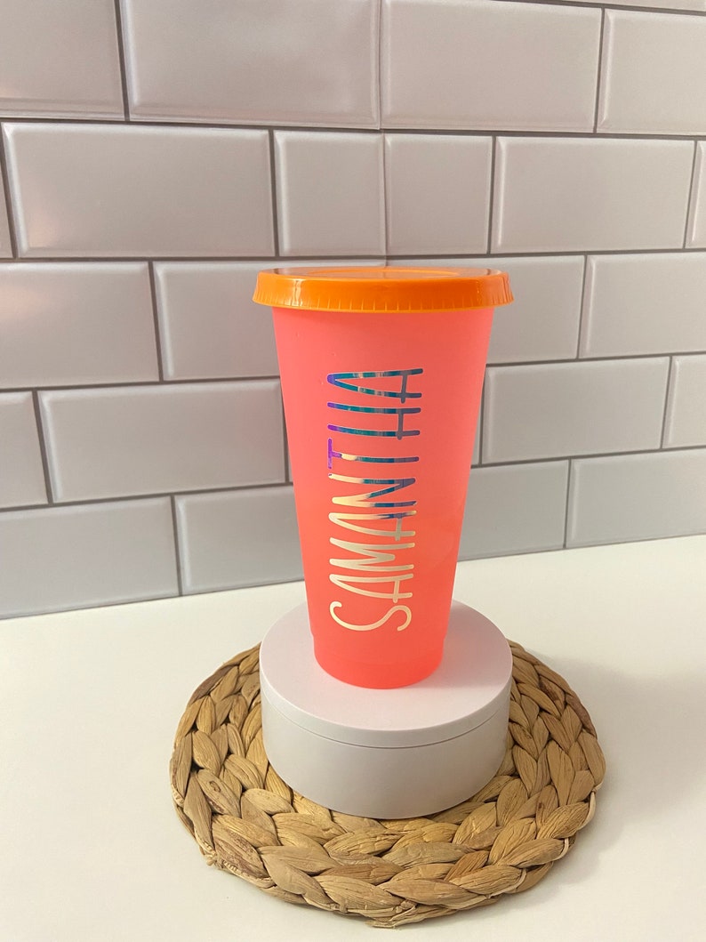 May include: A pink reusable cup with an orange lid and the name "Samantha" written on the side in a silver holographic font. The cup is sitting on a woven straw coaster.