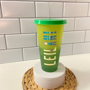 May include: A green and yellow ombre reusable cup with a green lid. The cup has a holographic sticker with the word "LEILA" on it.