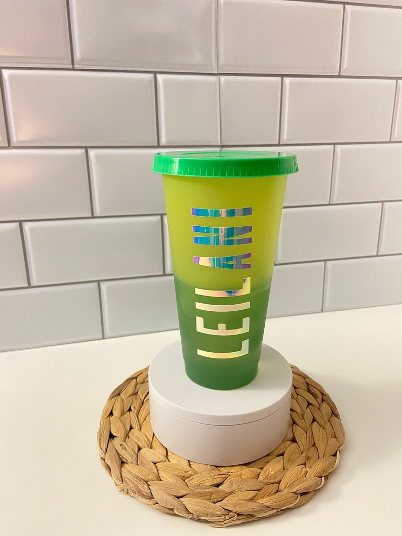 May include: A green and yellow ombre reusable cup with a green lid. The cup has a holographic sticker with the word "LEILA" on it.