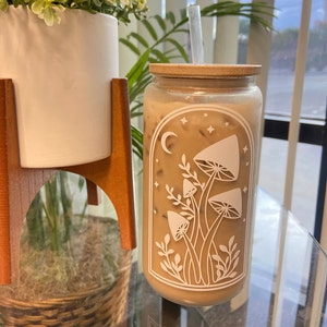 May include: A clear glass jar with a bamboo lid and a straw, filled with a light brown beverage. The jar has a white design of mushrooms and a crescent moon with stars.