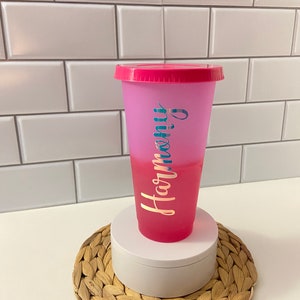 May include: A pink and purple ombre reusable cup with a pink lid. The cup has the word "Harmony" written on it in gold lettering.
