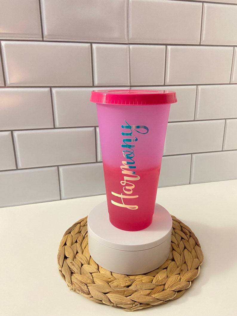 May include: A pink and purple ombre reusable cup with a pink lid. The cup has the word "Harmony" written on it in gold lettering.