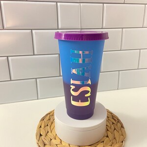May include: A blue and purple ombre reusable cup with a purple lid. The cup has the name "Isaiah" written on it in iridescent letters.