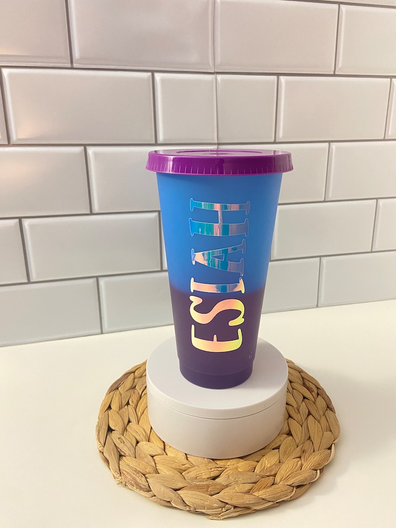 May include: A blue and purple ombre reusable cup with a purple lid. The cup has the name "Isaiah" written on it in iridescent letters.