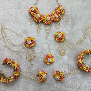 以下が含まれることがあります： 黄色とオレンジの花、パール、金色のアクセントが付いた花のジュエリーセット。セットにはネックレス、イヤリング、マングティカ、バングルが含まれています。