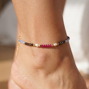 Peut inclure: Une chevillère composée d'une alternance de perles bleu clair, marron foncé et rose vif, agrémentée de petites perles blanches et d'entretoises en métal doré. Le bijou comporte une chaînette d'extension dorée avec un fermoir mousqueton.