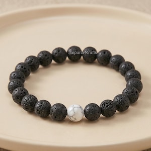 Op de afbeelding: Een zwarte lavasteen kralenarmband met één witte howliet kraal. De armband bestaat uit ronde, poreuze zwarte kralen en één witte kraal. De armband wordt gepresenteerd op een beige bord.