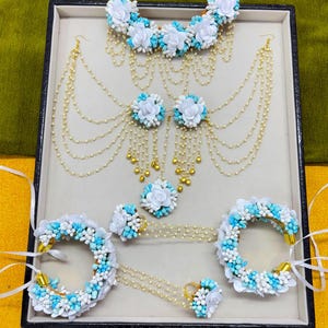 以下が含まれることがあります： 白と水色の花のアクセントが特徴的なブライダルジュエリーセット。セットには、ネックレス、イヤリング、指輪、ブレスレットが含まれており、すべて繊細なパールのストランドと金色のディテールが施されています。ジュエリーはニュートラルなトーンの表面に配置されています。