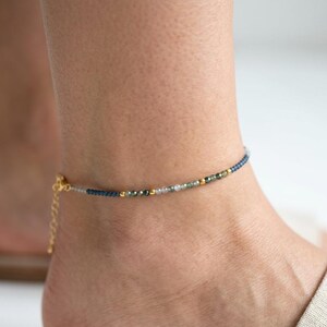 May include: A delicate anklet featuring a strand of small, faceted beads in shades of blue, green, and grey, interspersed with tiny gold-toned metallic spheres. The piece includes a gold-toned chain extender with a lobster claw clasp closure.