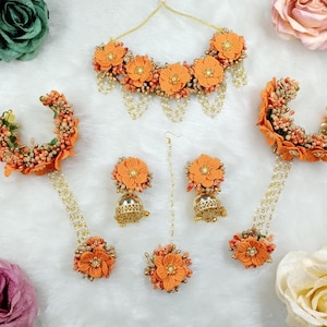 以下が含まれることがあります： オレンジ色の花のジュエリーセット。ネックレス、イヤリング、ヘアアクセサリー、ブレスレットが含まれています。セットには、金色のアクセントとぶら下がっているチェーンが付いたオレンジ色の布の花が含まれています。イヤリングはベル型です。
