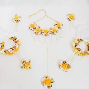 以下が含まれることがあります： ネックレス、イヤリング、バングル、マング・ティカなど、黄色と白の花のジュエリーセット。ジュエリーは造花とパールで作られています。
