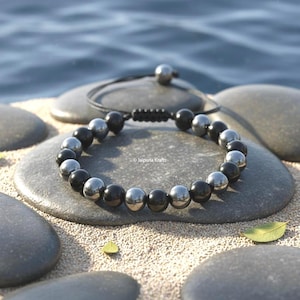May include: A bracelet with alternating black and silver beads. The beads are round. The bracelet is on a grey stone, with a black cord and a silver bead closure. The background includes sand, stones, and water.