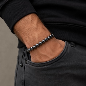 May include: A dark gray beaded bracelet. The bracelet is made of round, polished beads strung together. The bracelet is worn on a wrist, with the hand in a pocket. The person is wearing a black long-sleeve shirt and dark gray jeans.