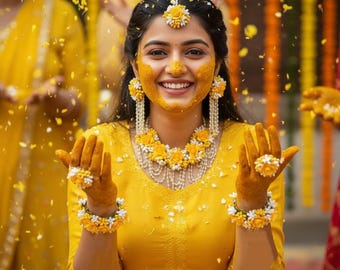 May include: A woman smiles, covered in yellow powder, wearing a yellow dress and floral jewellery. The jewellery includes a headpiece, earrings, a necklace, a bracelet, and a ring. Yellow flower petals fall around her.