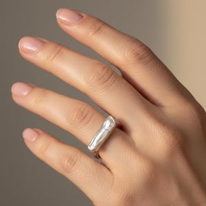 Puede incluir: Un anillo de plata con un diseño rectangular texturizado. El anillo se lleva en un dedo, mostrando su forma única y acabado pulido. El diseño del anillo es una versión moderna de un estilo clásico.