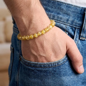 Könnte beinhalten: Ein goldgelbes Perlenarmband mit strukturierter Optik. Das Armband wird am Handgelenk getragen, wobei die Hand in einer blauen Jeans-Tasche ruht. Die Perlen scheinen halbtransparent zu sein.