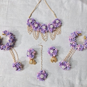May include: A purple and pink flower jewelry set with a necklace, earrings, a maang tikka, and two bangles. The jewelry is made with artificial flowers and gold chains.