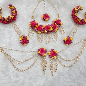 以下が含まれることがあります： ピンクと黄色の花のジュエリーセット。ネックレス、イヤリング、バングル、マングティカが含まれています。このセットは、造花とパールで作られています。