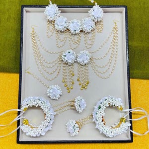 以下が含まれることがあります： 白い花のジュエリーセット。ネックレス、イヤリング、ヘッドピース、ブレスレット、ハンドジュエリーが含まれています。白い花のアクセントと真珠のようなビーズが特徴で、明るい灰色の表面に展示されています。