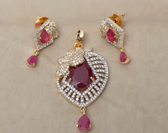 Può includere: Un set di collana con ciondolo in oro con una grande pietra rossa circondata da pietre di zircone cubico trasparenti. Il ciondolo ha la forma di un cuore con una pietra a forma di lacrima che pende dalla parte inferiore. Gli orecchini sono piccoli e presentano un'unica pietra rossa.
