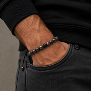 May include: A dark gray beaded bracelet with a mix of matte and metallic beads. The bracelet is worn on a wrist, with the hand in a pocket. The beads appear to be made of stone and metal.